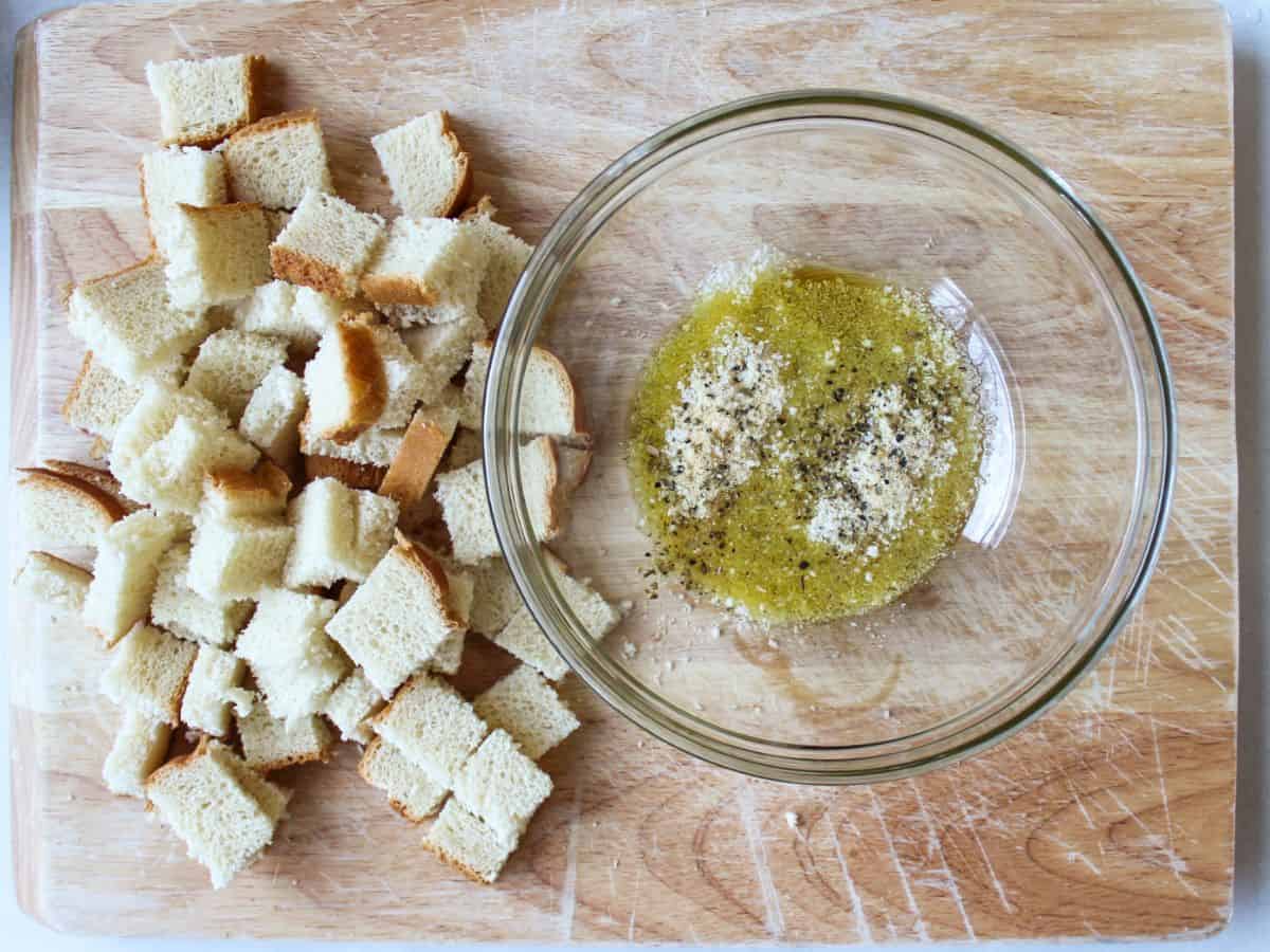 A cutting board with cubed fresh white bread on the left and a glass bowl with yellow oil and herbs on the right.