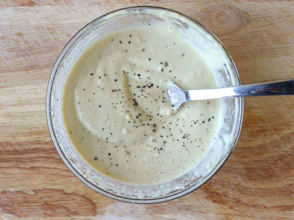 A glass bowl with creamy yellow dressing and a spoon in it. The bowl is on a wooden surface and there is specs of black pepper on top of the sauce.
