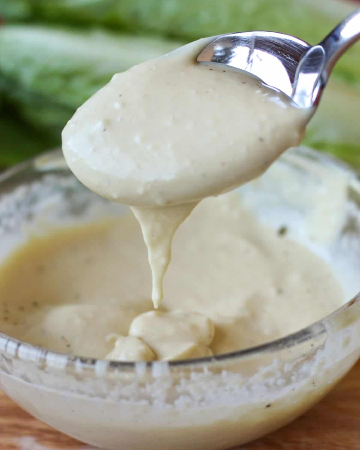 A glass bowl with creamy sauce on a wooden surface. The spoon filled with a sauce is dripping over the bowl. There is green lettuce in the background.