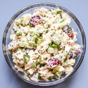 A glass bowl with creamy chicken salad garnished with thinly sliced green scallions. The background is light blue.