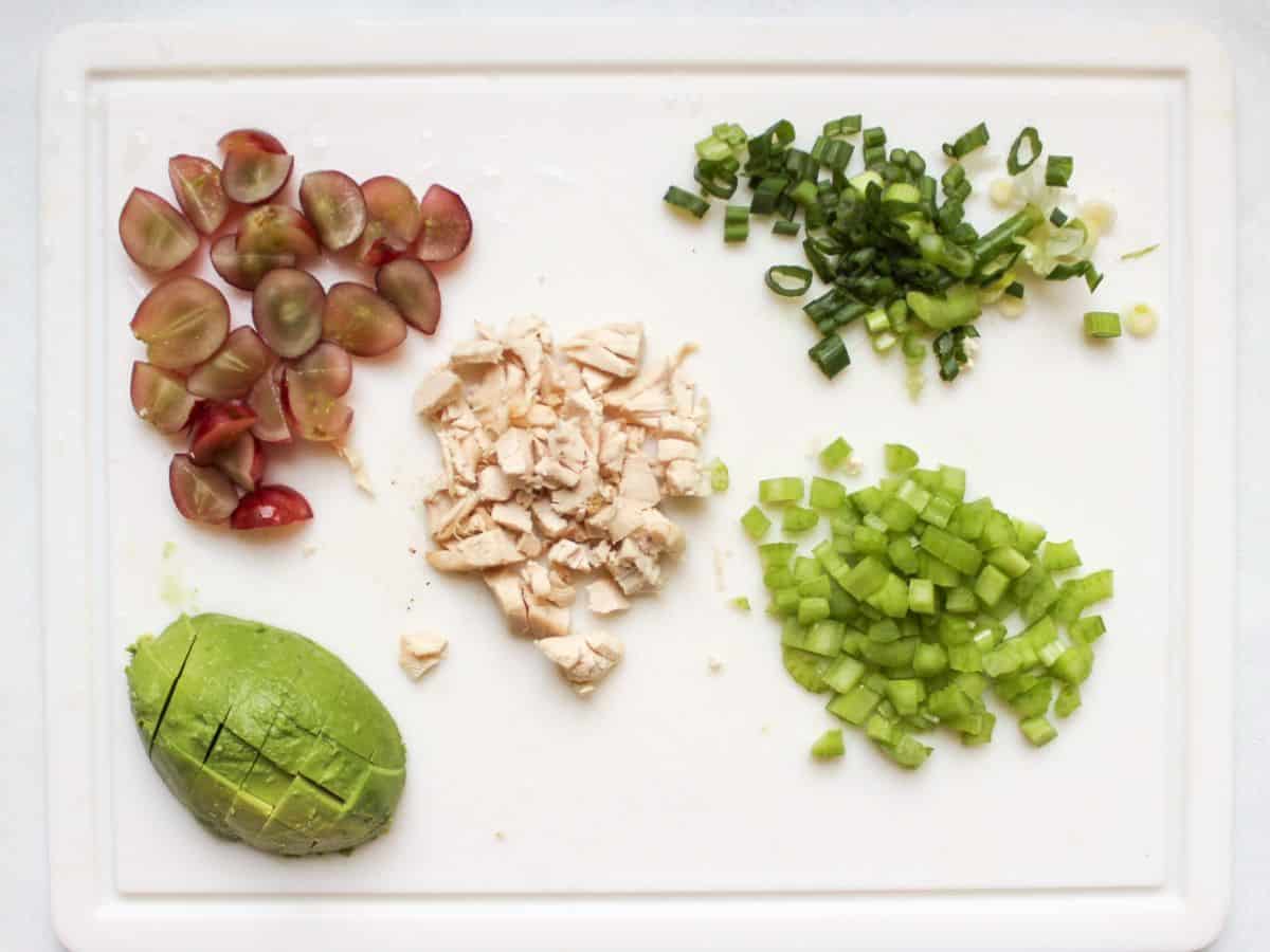 A white cutting board with quartered red grapes, diced chicken meat, diced green onions, diced green celery and cubed avocado.