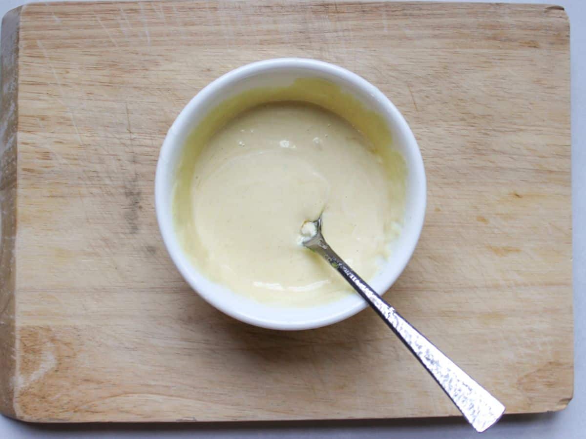 A small bowl with white creamy sauce and a spoon dipped in it. The bowl is on a wooden cutting board.
