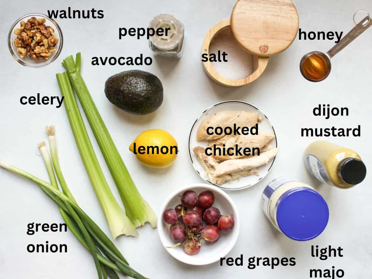 The labeled recipe ingredients on a white surface: pepper, whole avocado, salt, honey, walnuts, green whole scallions, two celery ribs, whole red grapes, a jar of mayo, Dijon mustard and two cooked chicken breasts.