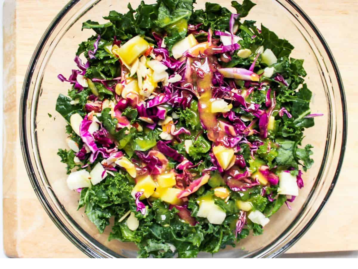 A glass bowl with chopped dark greens, purple shreds of cabbage, sliced almond and drizzles of yellow dressing on top.