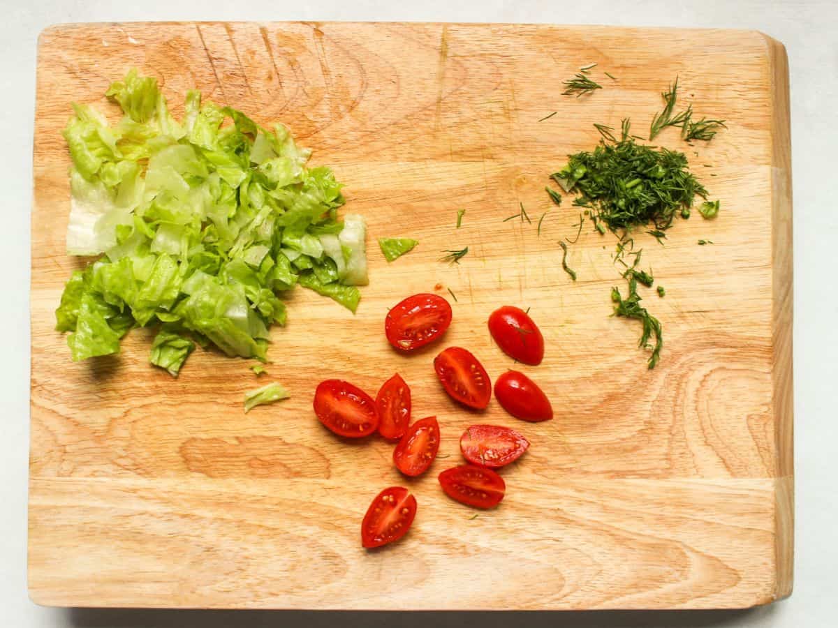 A cutting board with three piles of veggies: chopped lettuce, diced fresh dill, sliced in half cherry tomatoes.