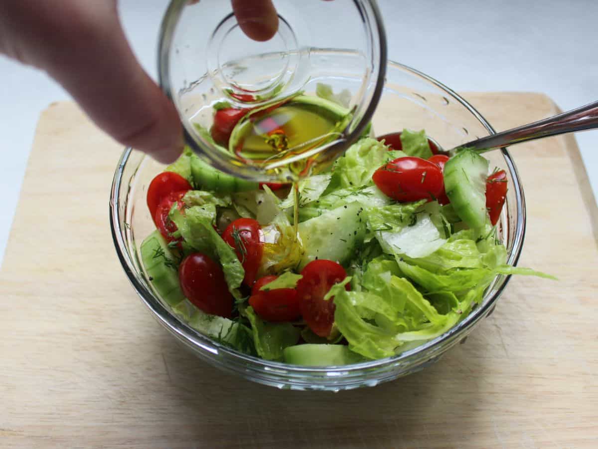 Olive oil is pouring from a small glass bowl into the lettuce, cucumber and tomatoes salad. The salad bowl is glass as well.