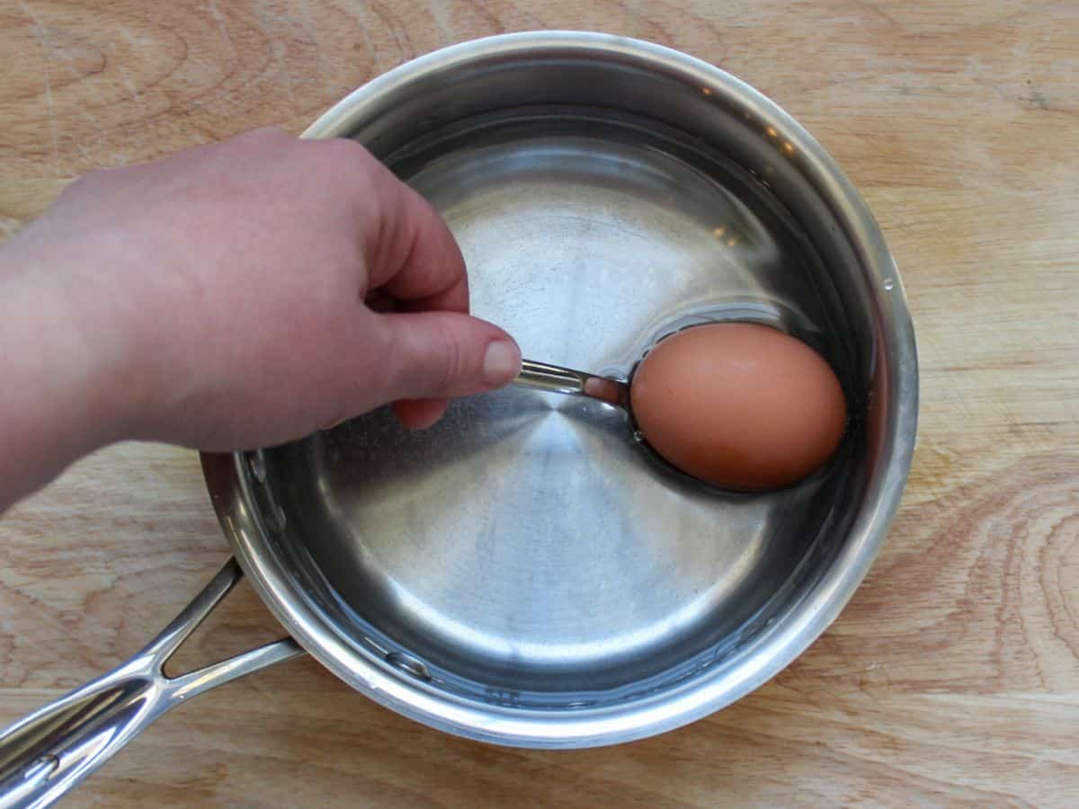 A spoon with an egg being submerged into a stainless steel saucer pan filled with water.