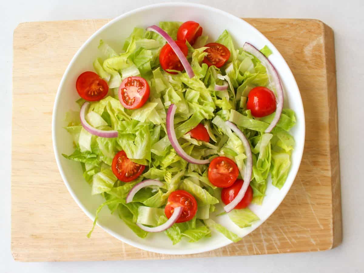 A shallow white dish with chopped lettuce, halved cherry tomatoes and sliced red onions on a wooden board.
