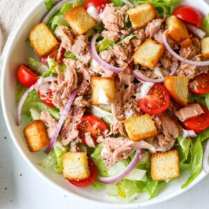 A white shallow dish with a salad made of chopped lettuce, halved cherry tomatoes, chunks of tune, sliced red onion and croutons.