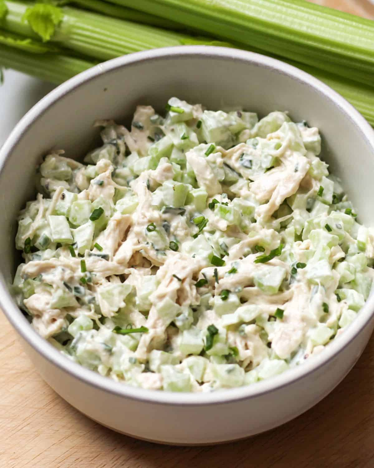 Creamy salad made with white shredded meat, diced green celery in a white bowl. There is a talk of green fresh celery behind the bowl.