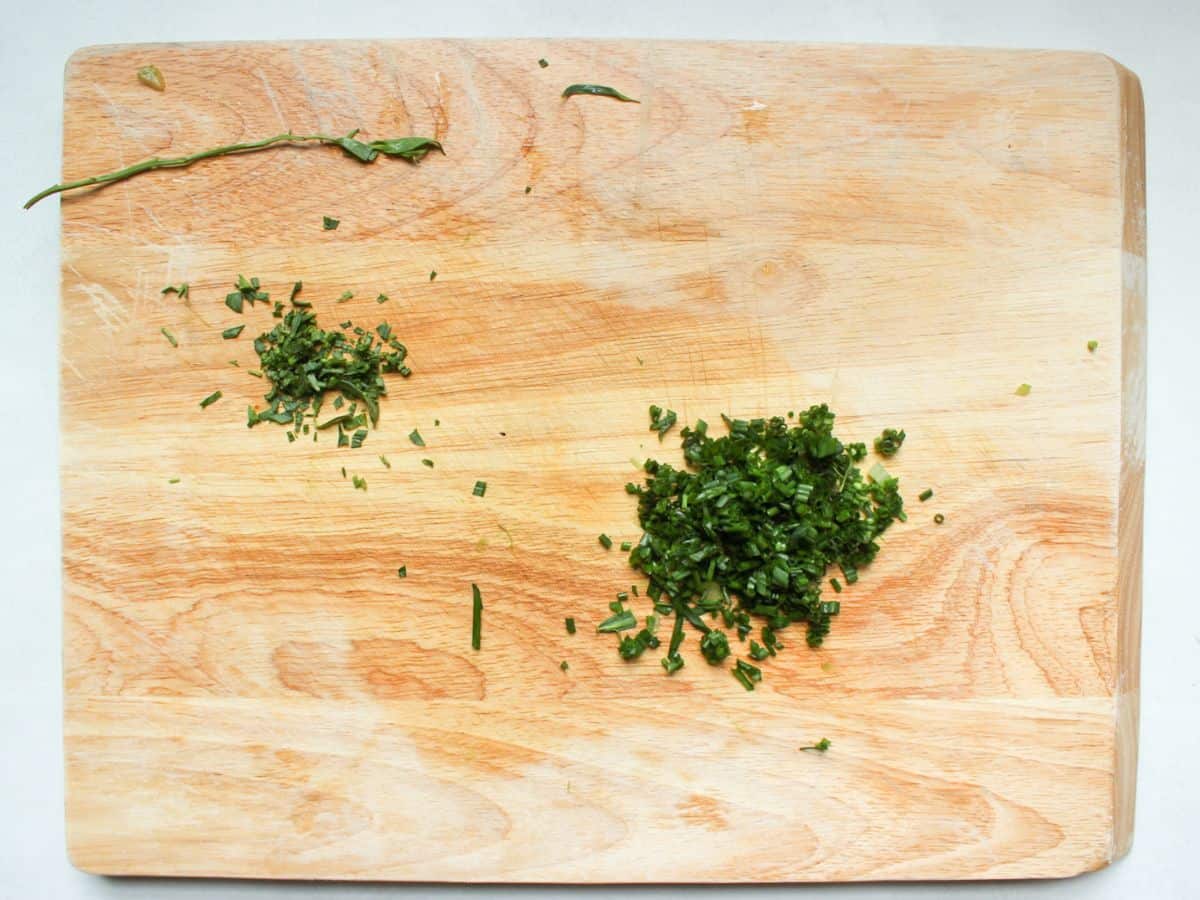 Two heaps of finely chopped green chives and tarragon on a wooden surface. There is also a green stem on the side.