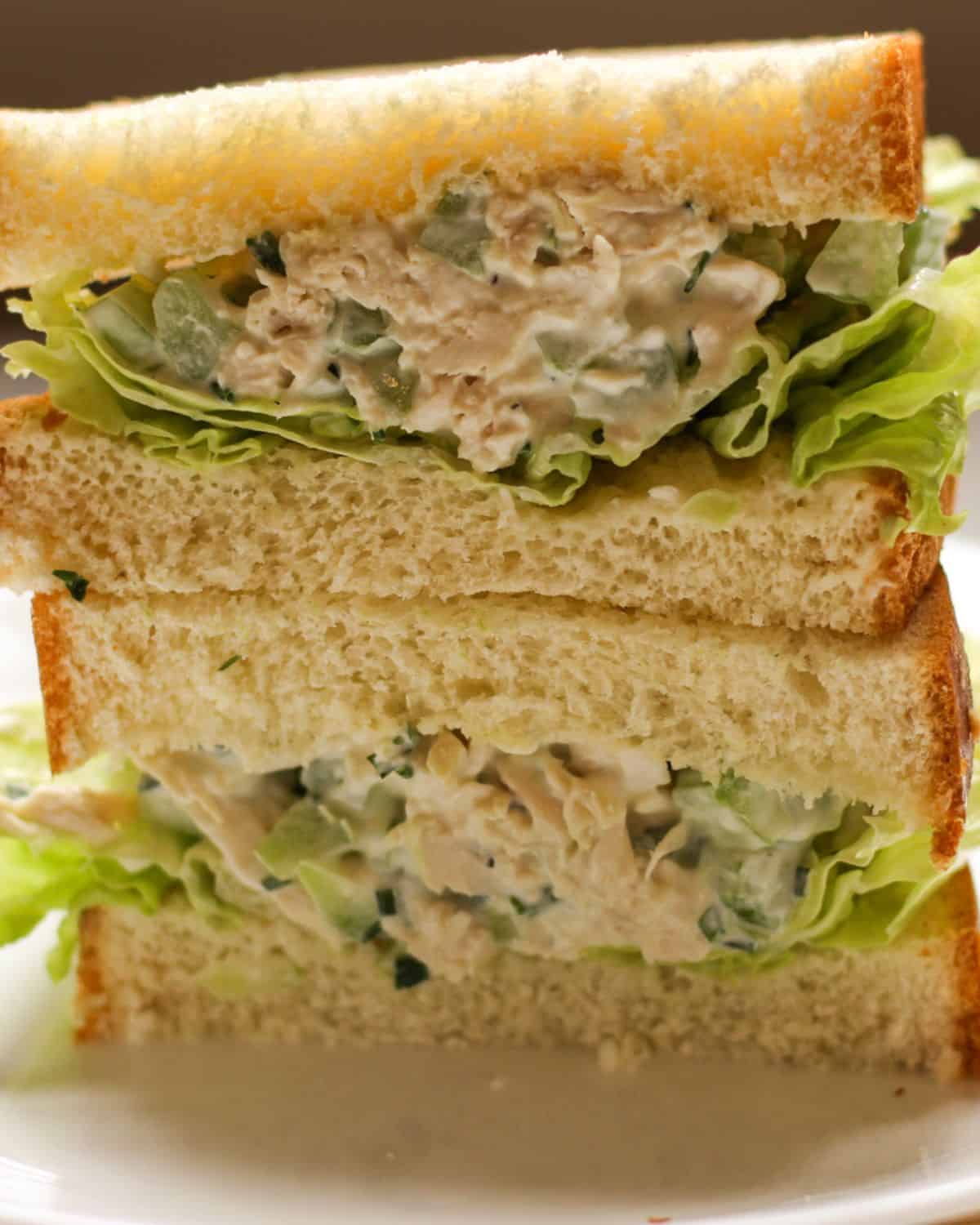 Two chicken sandwich halves stacked on top of each other. The sandwich is made with white bread, green lettuce and creamy chicken salad.