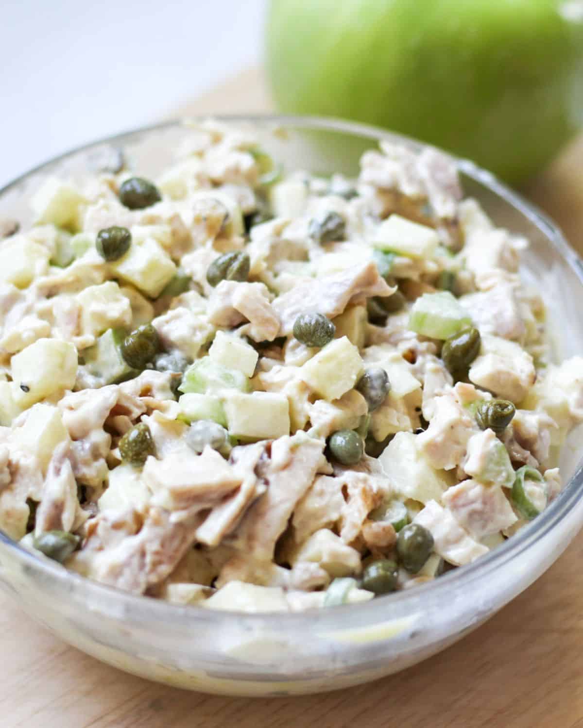 Creamy chicken salad with cubed apples and capers in a large glass bowl. There is a whole green apple behind the bowl.