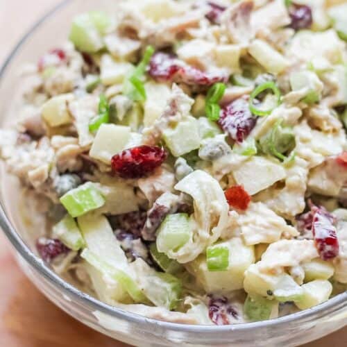 Large glass bowl, almost fully visible, filled with a creamy chicken salad featuring chopped ingredients and red dried cranberries, garnished with diced green onion on top