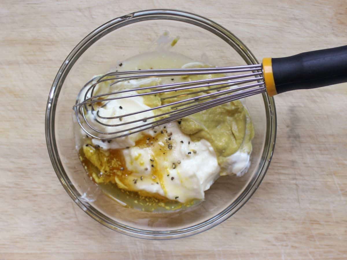 Small glass bowl with unmixed dressing ingredients, including white creamy sauce, mustard, lemon juice, liquid honey, and black pepper specks, with a whisk inside the bowl.