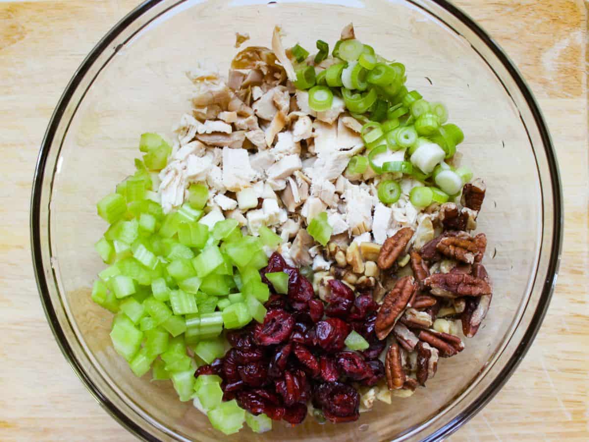 Large glass bowl containing unmixed chicken salad ingredients, including diced celery, shredded chicken, chopped pecans, diced green onions, and dried cranberries.