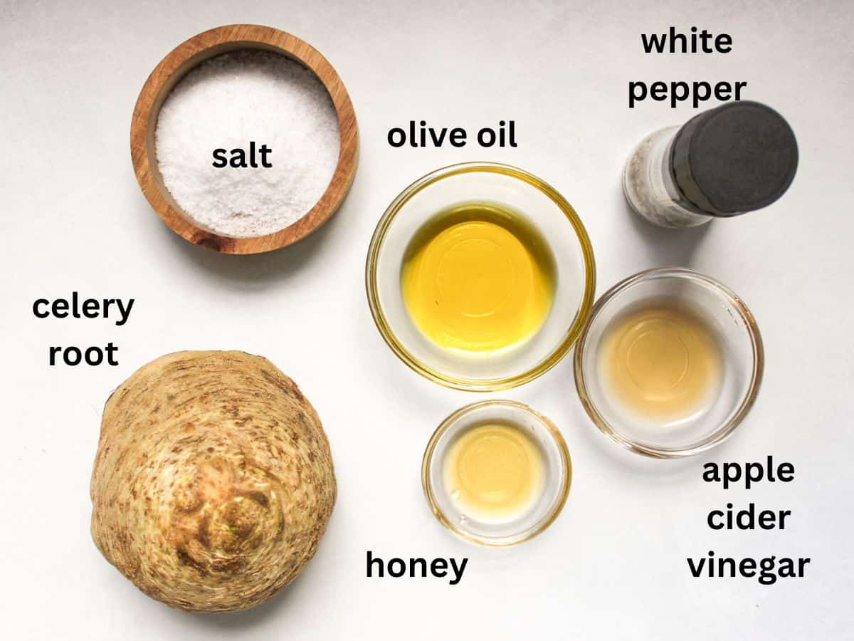 German celery root salad labeled ingredients on a white surface. From left to right: celery root, salt, olive oil, honey, apple cider vinegar, white pepper.