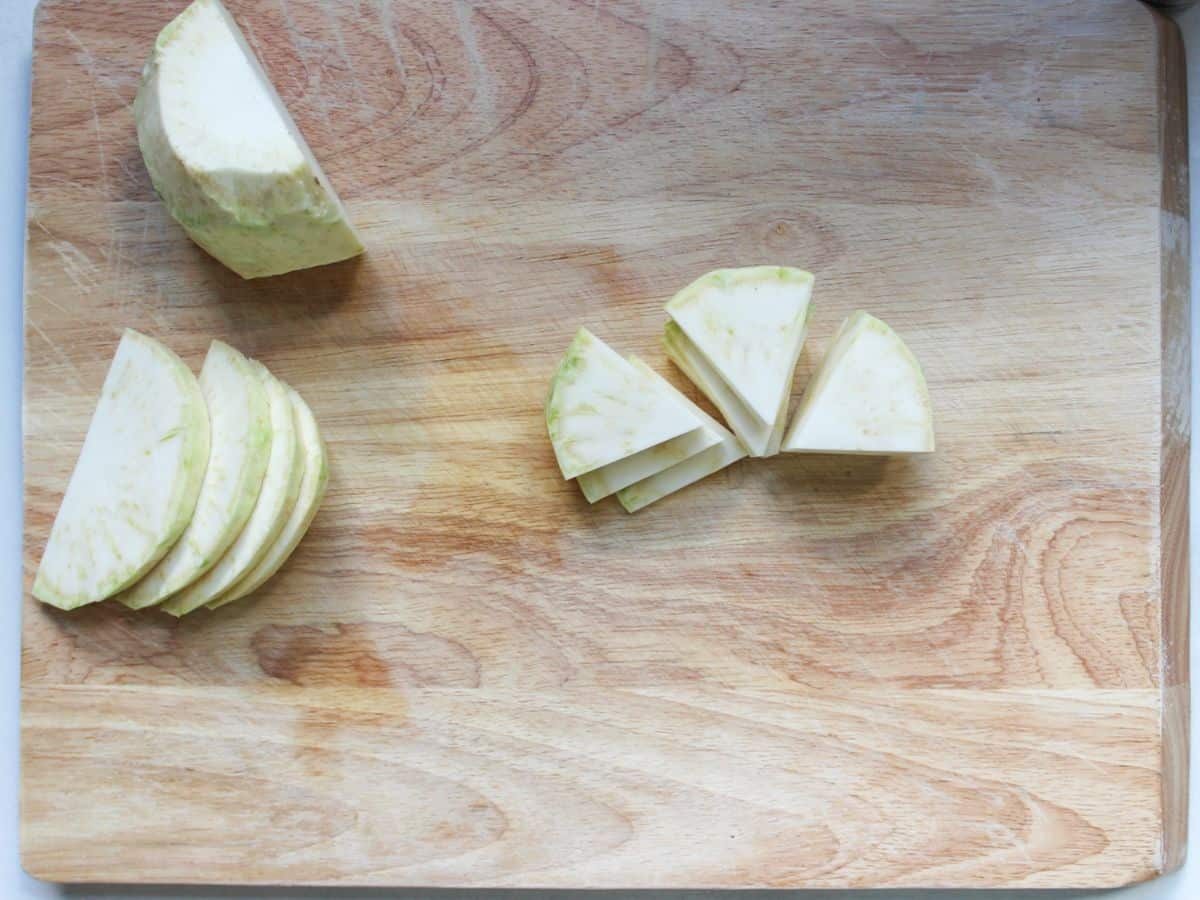A cutting board with half of the celery root, a stack of half-circle slices, and a stack of the half-circle slices being cut into three parts.