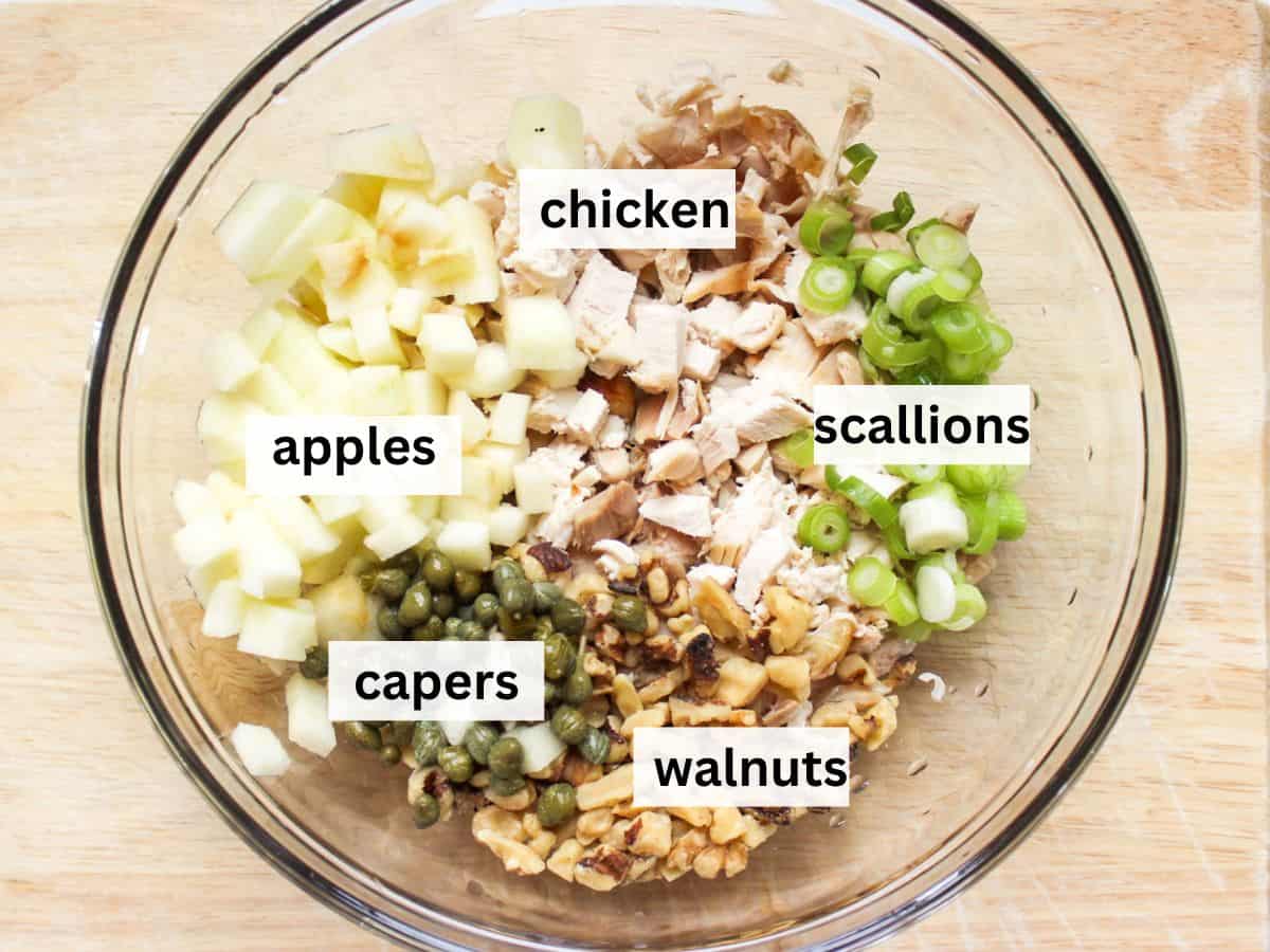 5 separate heaps of cubed apples, diced chicken, diced scallions, chopped walnuts and drained capers in a large glass bowl.
