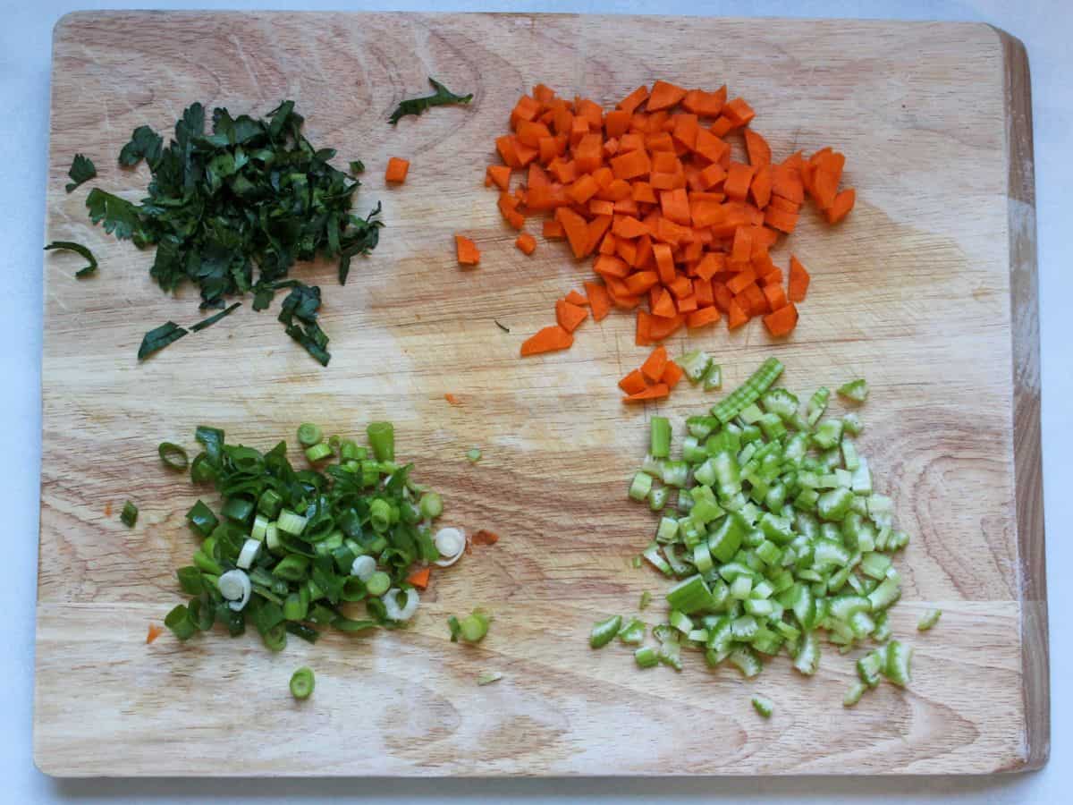 four separate heaps of chopped vegetables and herbs on a wooden cutting board: parsley, carrots, green onions, celery.