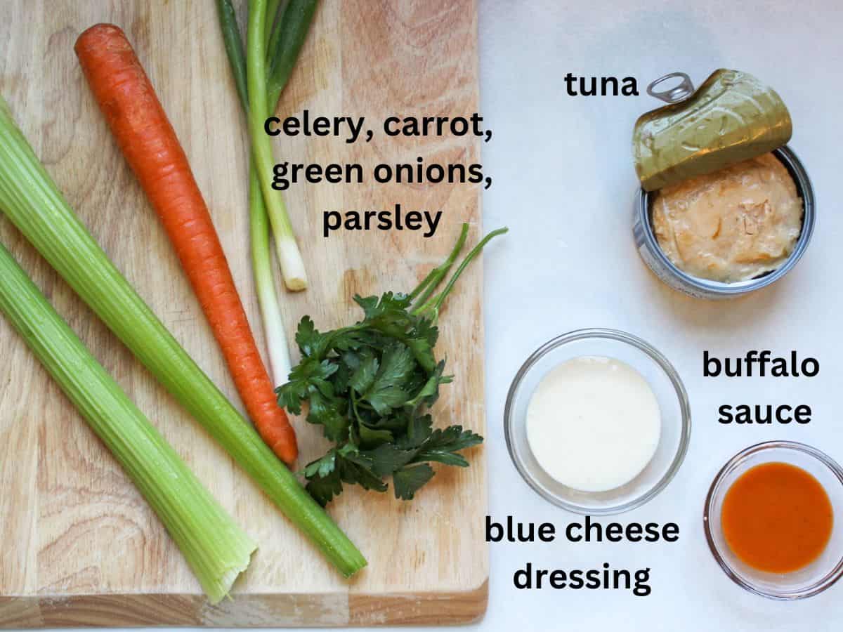 Recipe ingredients on a white cutting board and a white background: celery, carrot, green onion, parsley, blue cheese dressing, open can of tuna, red buffalo sauce.