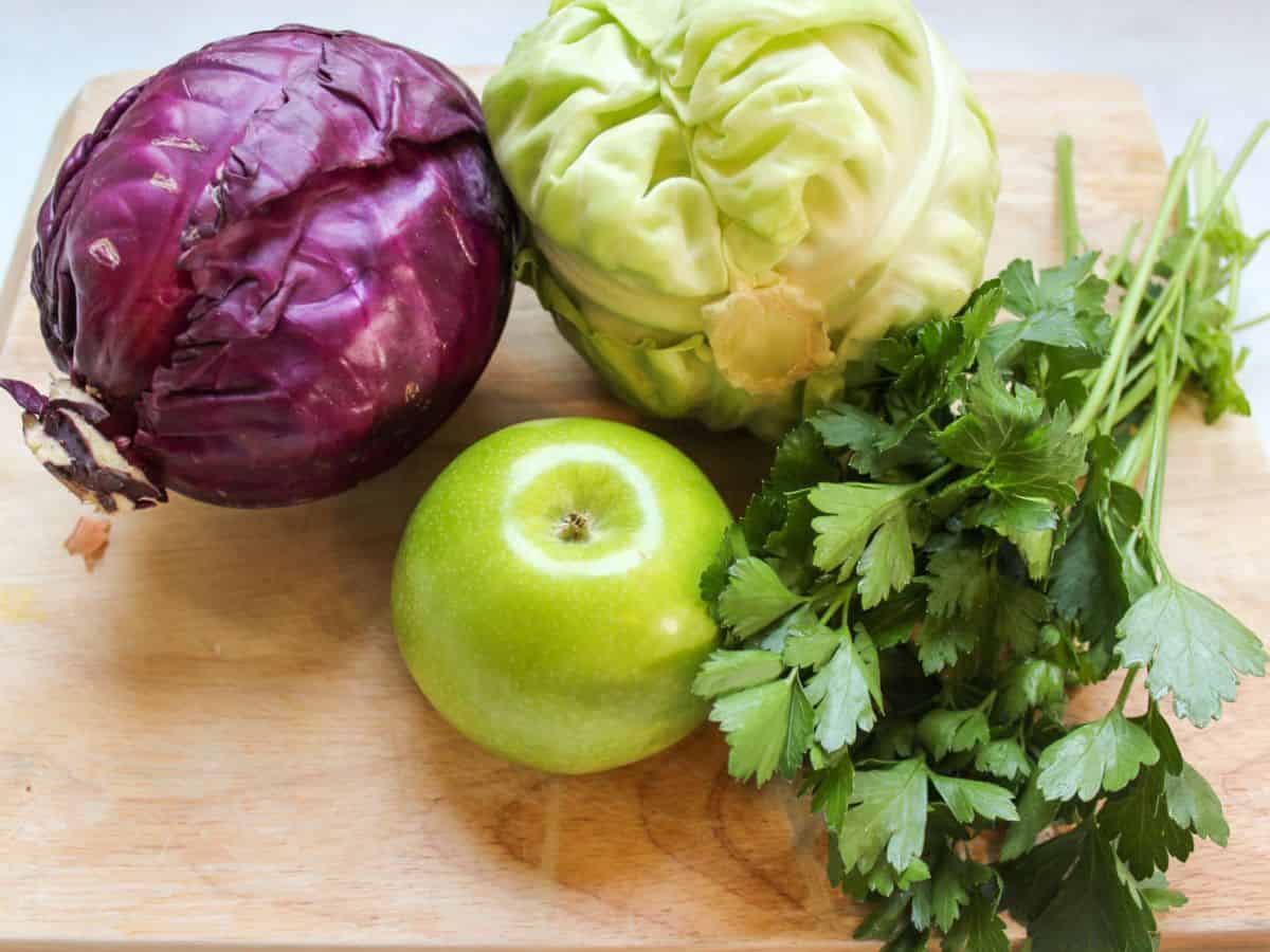 Whole red cabbage, whole green cabbage, whole green apple and a bunch of fresh parsley on a wooden cutting board.